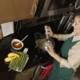 Mulher cozinhando em uma cozinha moderna, derramando um condimento sobre uma panela; ao seu lado há molho, aspargos e bananas sobre a bancada.