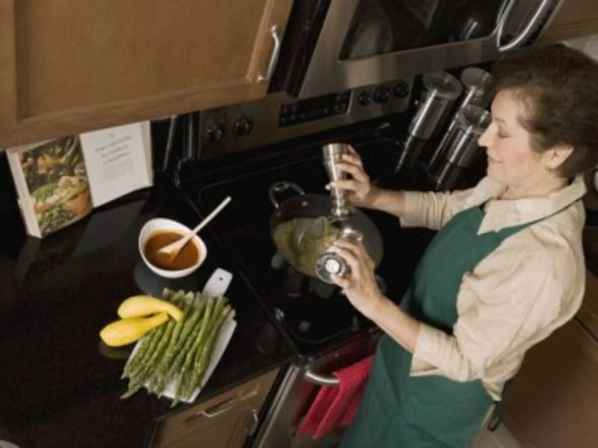Mulher cozinhando em uma cozinha moderna, derramando um condimento sobre uma panela; ao seu lado há molho, aspargos e banana-da-terra sobre a bancada.