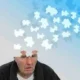 Homem idoso com expressão de preocupação, peças de um quebra-cabeça se dispersam sobre fundo azul, simbolizando a memória e a qualidade de vida na doença de Alzheimer.
