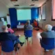 Grupo de pessoas em uma sessão de reabilitação cognitiva diante de uma tela com grade e um apresentador à frente.