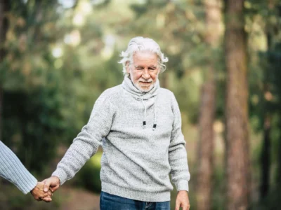 Idoso com cabelos grisalhos e barba curta sorri enquanto segura a mão de outra pessoa, diante de uma floresta com luz suave.