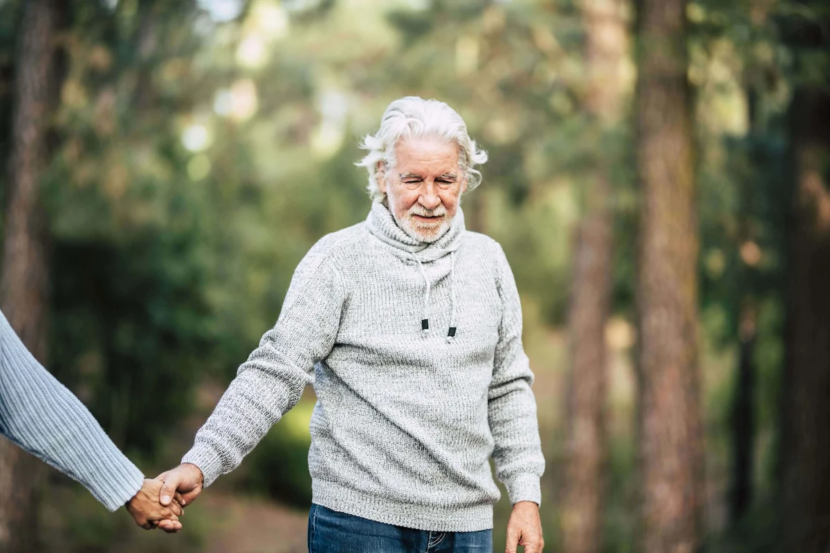 Homem idoso com cabelo grisalho e barba curta sorri enquanto segura a mão de outra pessoa, em frente a uma floresta com luz suave.