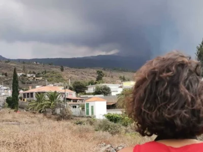 Pessoa de costas com cabelo cacheado observa uma paisagem de La Palma: casas, montanhas e céu nublado em um ambiente árido.