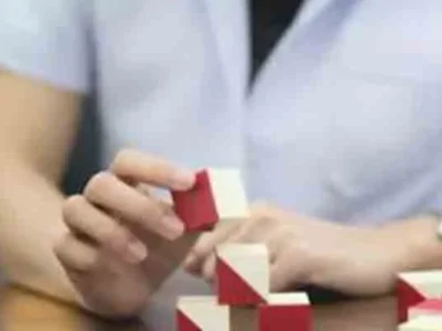 Mão segurando um bloco de brinquedo vermelho e branco enquanto se constrói uma estrutura, em um ambiente de sala de aula.