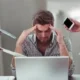 Profissional no escritório com expressão de estresse, mãos na cabeça, computador ligado e papéis sobre a mesa.