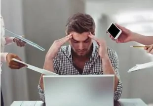 Profissional no escritório com expressão de estresse, mãos na cabeça, computador ligado e papéis sobre a mesa.