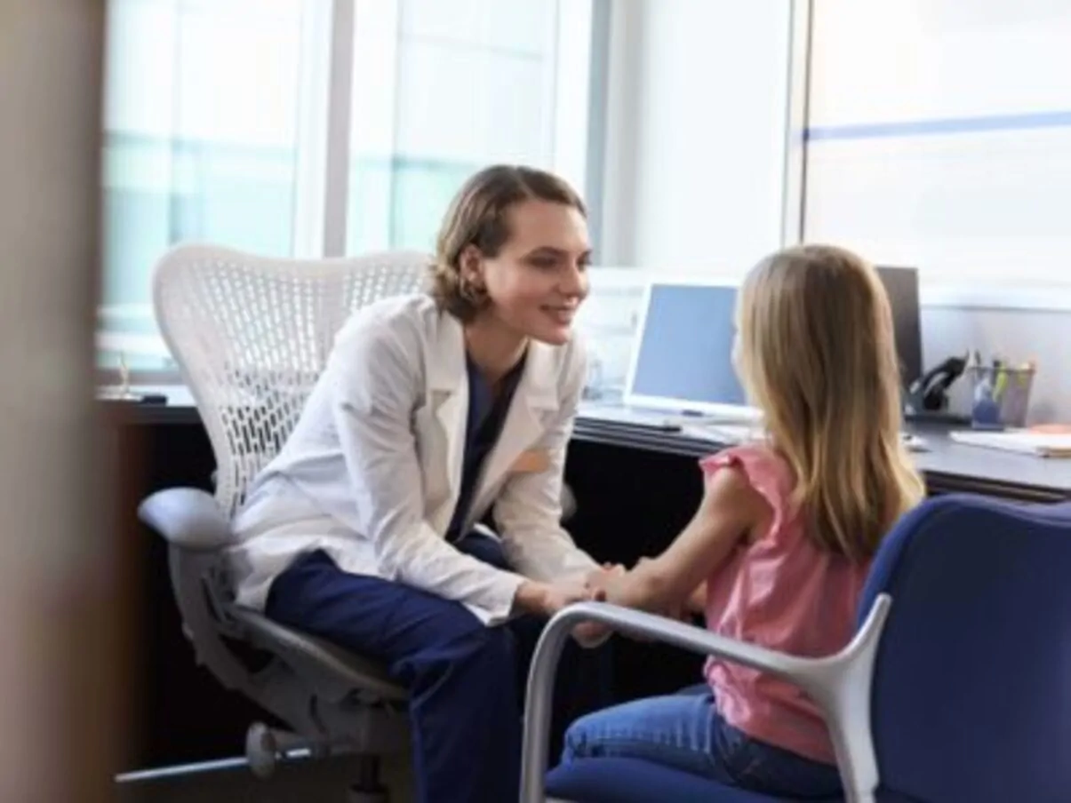 Pediatra conversa com uma menina em um consultório; bata branca e ambiente clínico que transmite atenção infantojuvenil.
