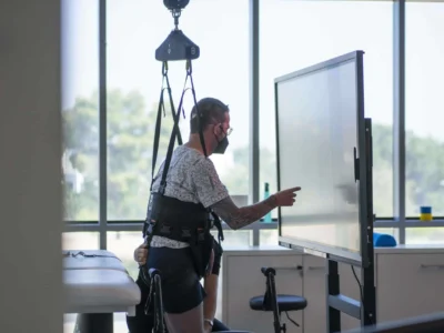 Pessoa em neuroreabilitação em pé com arnês interagindo com uma tela sensível ao toque, com iluminação natural
