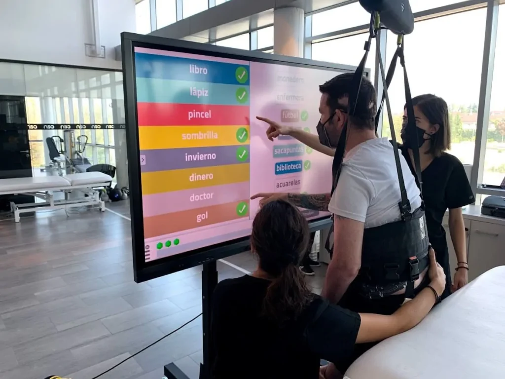 Paciente em arnês interage com tela sensível ao toque durante neurorreabilitação de dano cerebral, acompanhado por equipe.