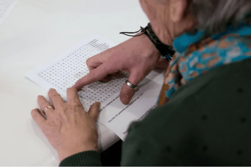 Mulher idosa com Parkinson participa de fonoaudiologia para linguagem; uma mão jovem aponta uma palavra em uma sopa de letras.