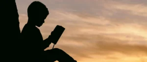 Silhueta de uma criança sentada segurando um livro diante de um entardecer quente, céu laranja e uma figura escura lendo.
