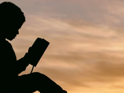 Silhueta de uma criança sentada segurando um livro em frente a um pôr do sol quente, céu laranja e figura escura lendo.