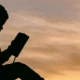 Silhueta de uma criança sentada segurando um livro em frente a um pôr do sol quente, céu laranja e figura escura lendo.