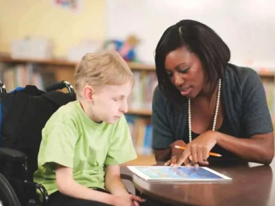 A terapeuta ocupacional ensina uma criança em cadeira de rodas usando um tablet, em um ambiente escolar.