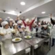 Grupo de pessoas com síndrome de Down participa de uma aula de culinária, com toques de chef e aventais brancos, em um ambiente alegre.