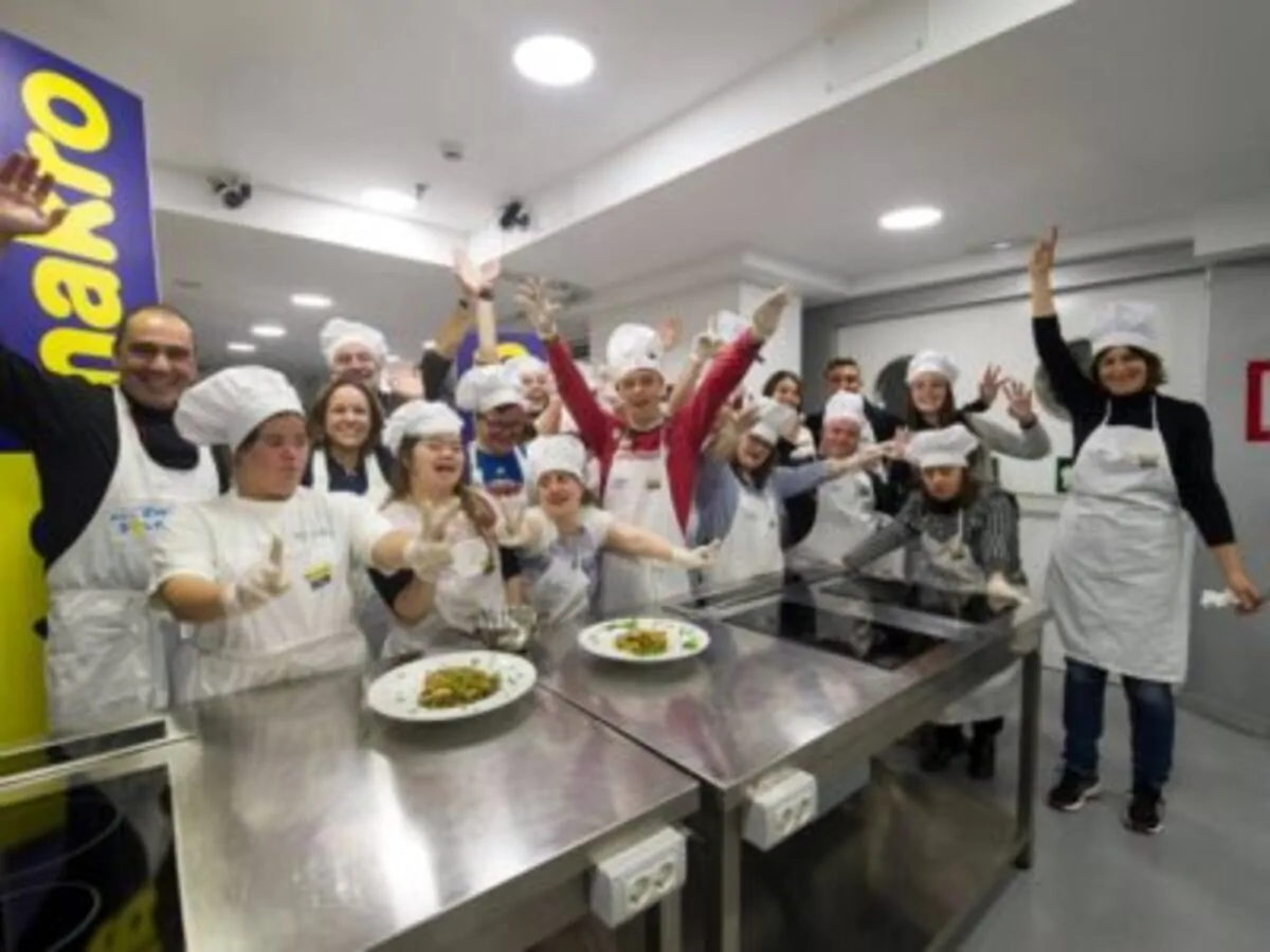 Grupo de pessoas com Síndrome de Down participa de uma aula de culinária, com toucas de chef e aventais brancos, em um ambiente alegre.