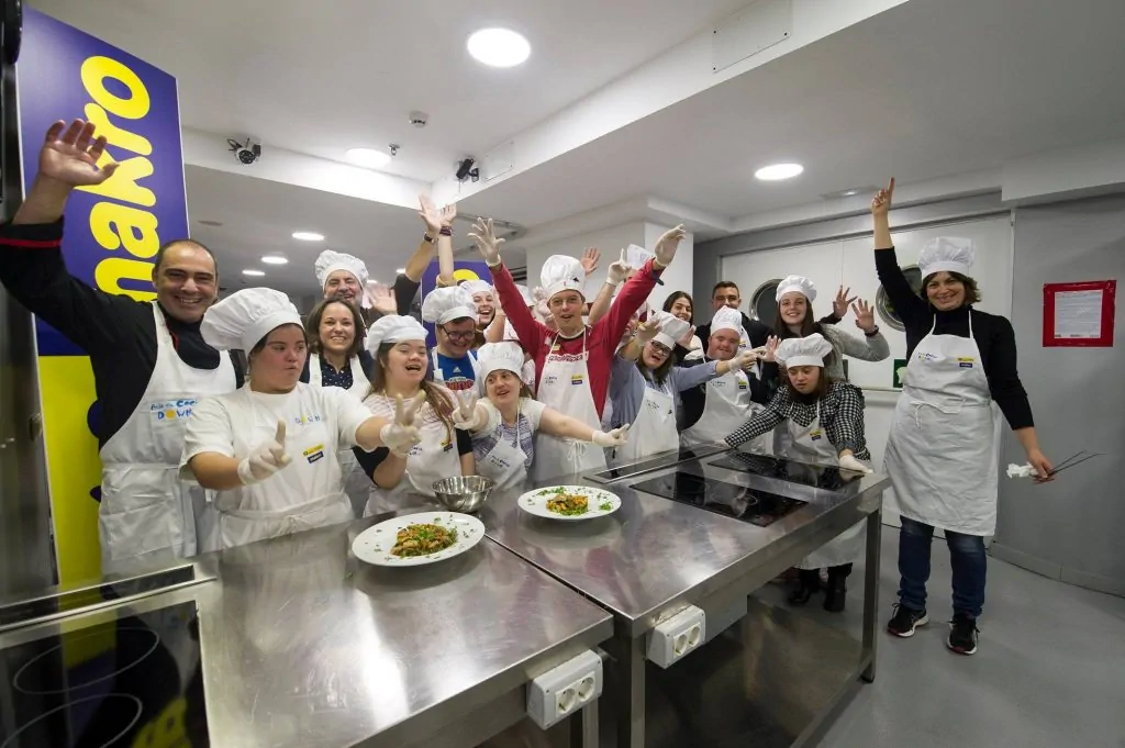 Grupo de pessoas com síndrome de Down em uma cozinha, com aventais e toucas de chef, celebrando junto a uma mesa de trabalho.