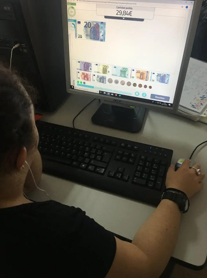 Persona frente a una computadora con auriculares, interactuando con una interfaz educativa que muestra billetes y euros.