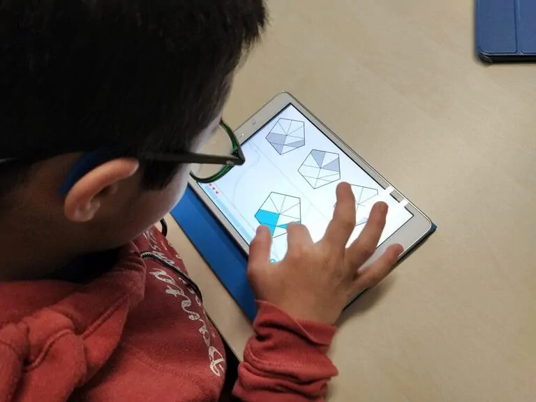 Menino com óculos e fones de ouvido usa um tablet em uma sala de aula, mostrando figuras geométricas enquanto participa de uma atividade educativa.