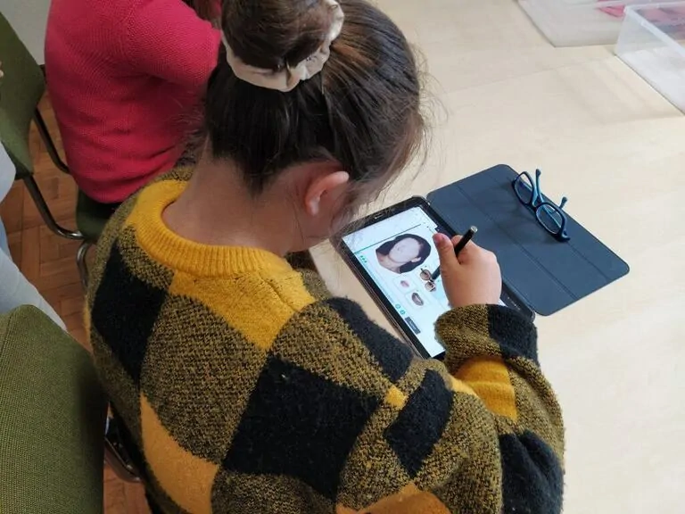 Menina sentada em uma cadeira usa um tablet com caneta digital para desenhar, vestindo um suéter xadrez amarelo e preto.