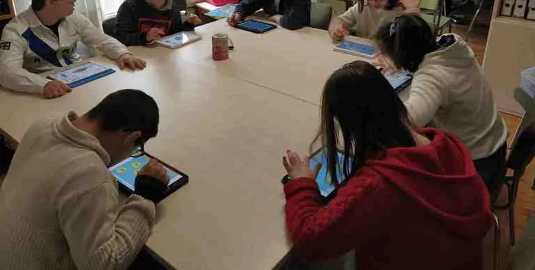 Grupo de pessoas com síndrome de Down ao redor de uma mesa redonda, com tablets, participando de aprendizagem cognitiva.