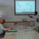 Grupo de participantes em sala de reuniões observando uma apresentação; uma pessoa em pé aponta para a tela, com máscaras e divisórias.