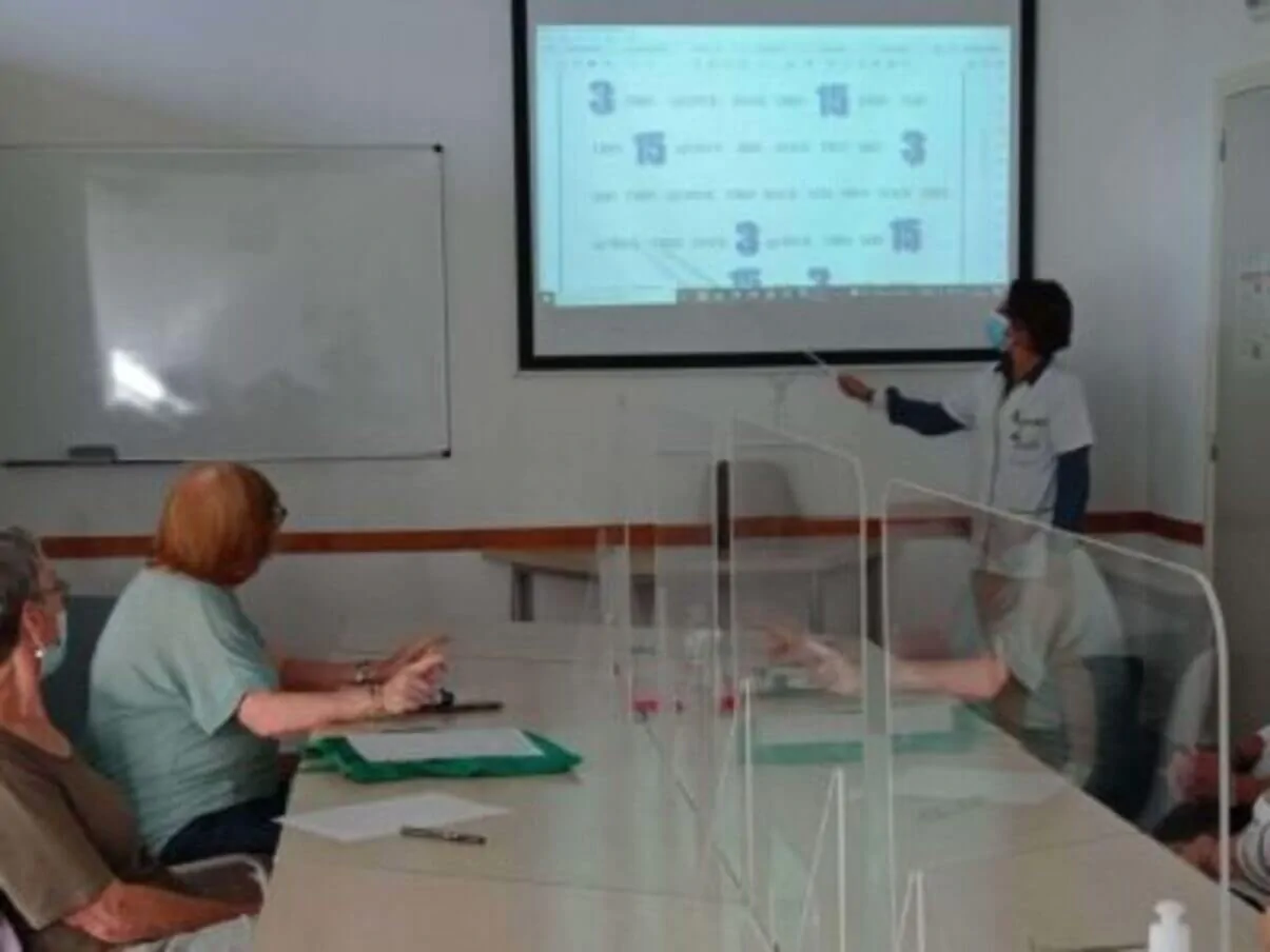 Grupo de participantes em sala de reuniões observando uma apresentação; uma pessoa em pé aponta para a tela, com máscaras e barreiras de proteção.