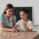 Uma mulher e uma menina sentadas diante de uma mesa, brincando com peças em uma atividade educativa, com uma lousa ao fundo.