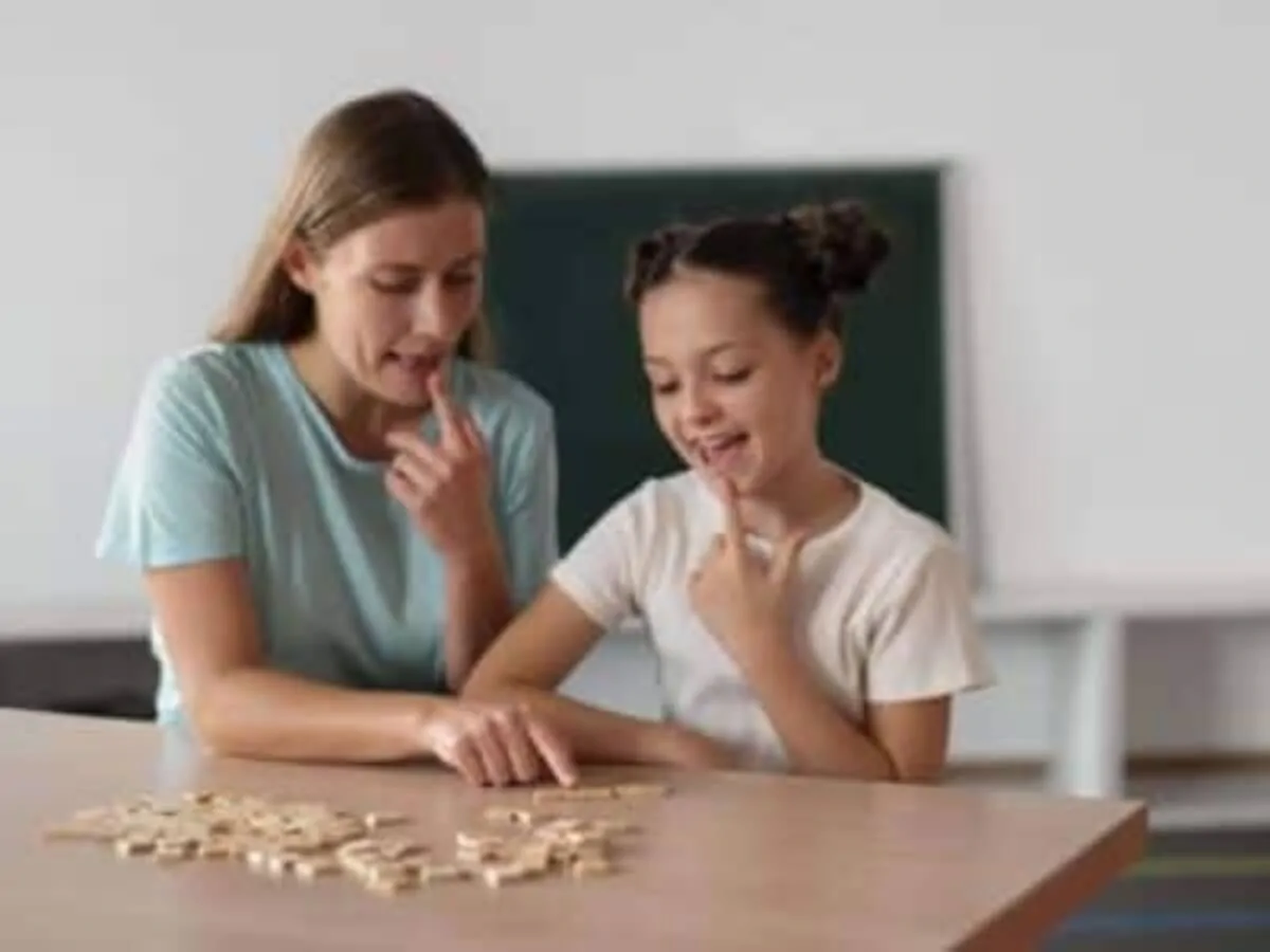 Uma mulher e uma menina sentadas em frente a uma mesa, brincando com peças em uma atividade educativa, com uma lousa ao fundo.