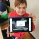 Criança sorridente com moletom vermelho segura um tablet em uma sala de aula; faz parte de um grupo de pessoas com síndrome de Down, ambiente de aprendizagem.