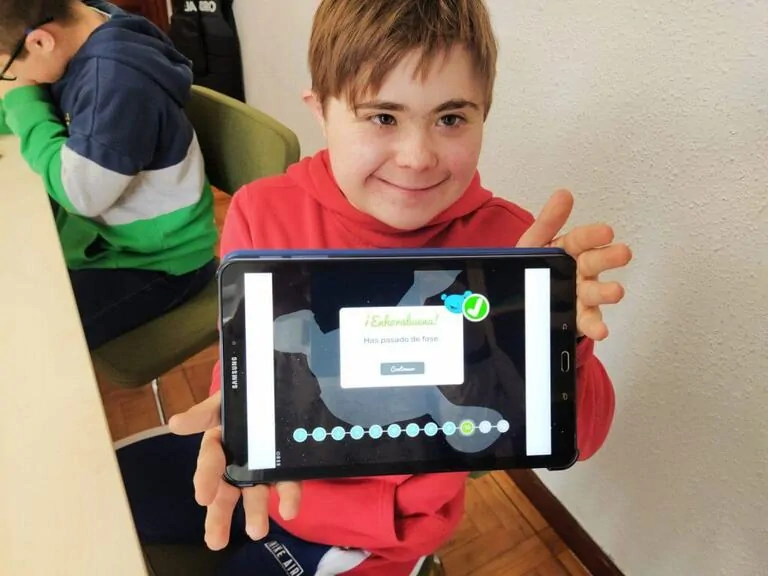 Menino sorridente com moletom vermelho segura um tablet em uma sala de aula; a tela mostra '¡Excelente!' enquanto outra pessoa observa ao fundo.