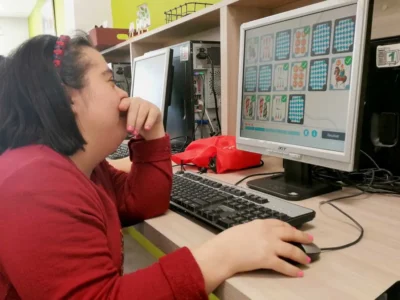 Menina com cabelo escuro sentada diante de um computador em uma sala de aula colorida, concentrada em uma atividade educativa.