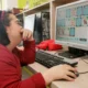 Menina com cabelo escuro sentada diante de um computador em uma sala de aula colorida, concentrada em uma atividade educativa.