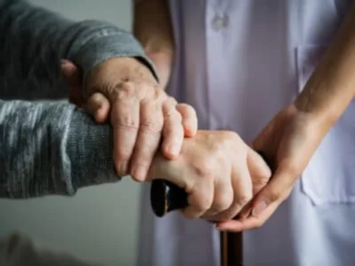 Primeiro plano de duas mãos entrelaçadas: uma pessoa idosa com bengala recebe apoio de um cuidador, em um ambiente de cuidado.