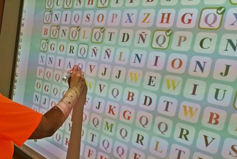 Tela interativa da NeuronUP com uma grade de letras coloridas e uma mão apontando uma célula.