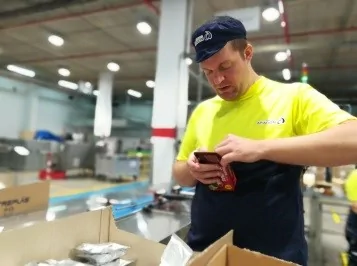 Homem num ambiente produtivo de produção com camiseta amarela e boné, segurando um celular e revisando uma caixa com pacotes.
