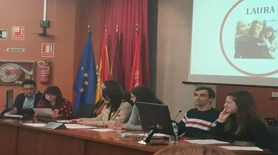 Painel de pessoas em um evento: apresentador com microfone, tela com o nome 'LAURA' e bandeiras, em ambiente acadêmico.