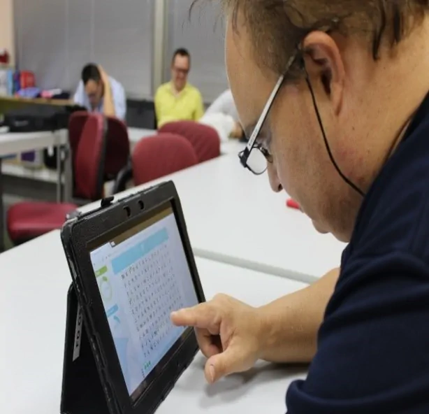 Homem ao lado de uma mesa com tablet; ao fundo, outras pessoas em uma sessão de estimulação cognitiva em uma sala.