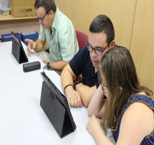 Três pessoas com síndrome de Down ao redor de uma mesa, concentradas em um tablet durante uma sessão de estimulação cognitiva.