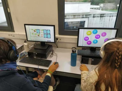 Dois meninos em uma sala de aula com fones de ouvido participam da telereabilitação com o NeuronUP em frente aos computadores.