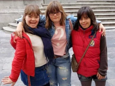 Três jovens posam ao ar livre: uma com jaqueta vermelha e saia, outra com camiseta clara e jeans, a terceira com jaqueta vermelha.