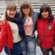 Três jovens posam ao ar livre: uma com jaqueta vermelha e saia, outra com camiseta clara e jeans, a terceira com jaqueta vermelha.