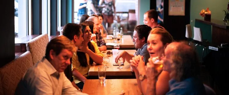 Grupo de pessoas sentadas ao redor de uma mesa em um bar ou restaurante, trocando ideias e gestos.
