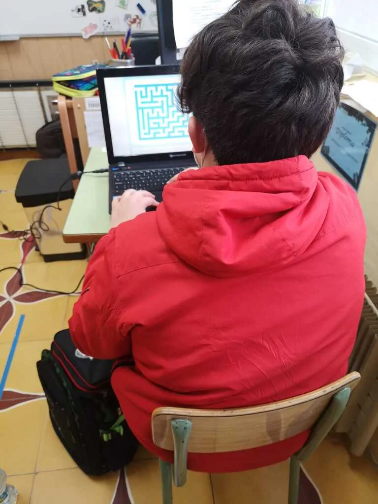 Aluno em sala com NeuronUP em notebook, concentrado em um labirinto; ambiente de sala de aula com quadro, mobiliário e mochila próxima.
