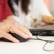 Mão de pessoa idosa usando o mouse; ao fundo, outra mão com o teclado, em uma aprendizagem centrada na estimulação cognitiva.