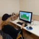 Mulher loira com boné preto sentada em frente a um computador; a tela mostra um jogo simples e uma paisagem, com uma bolsa e uma caneta na mesa.
