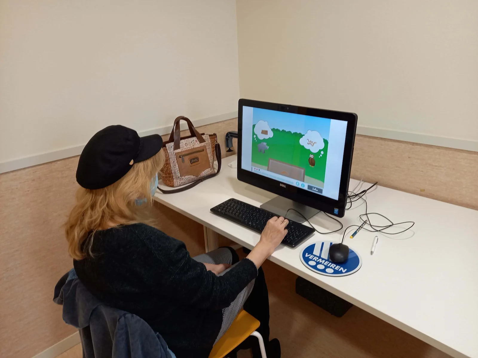 Mulher loira com boné preto sentada em frente a um computador; a tela mostra um jogo simples e uma paisagem, com uma bolsa e uma caneta na mesa.