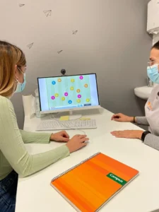 Duas pessoas com máscaras diante de um computador; tela com gráficos e círculos numerados, caderno laranja e garrafa.