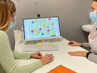 Duas pessoas com máscara em frente a um computador; tela com gráficos e círculos numerados, caderno laranja e garrafa.