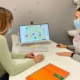 Duas pessoas com máscara em frente a um computador; tela com gráficos e círculos numerados, caderno laranja e garrafa.